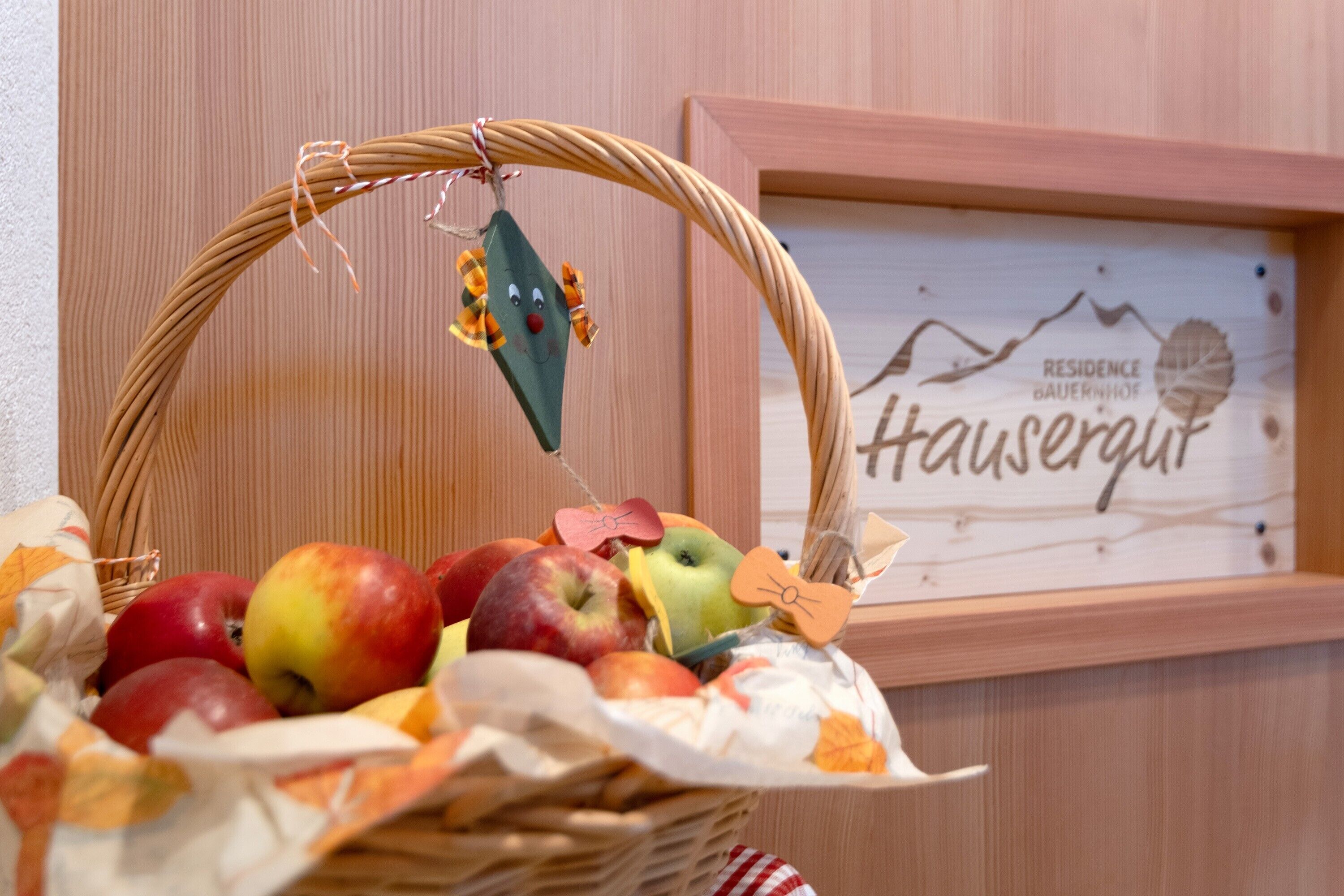 A wicker basket filled with apples and decorative items, placed in front of a framed sign with the text 'Hausergut.'