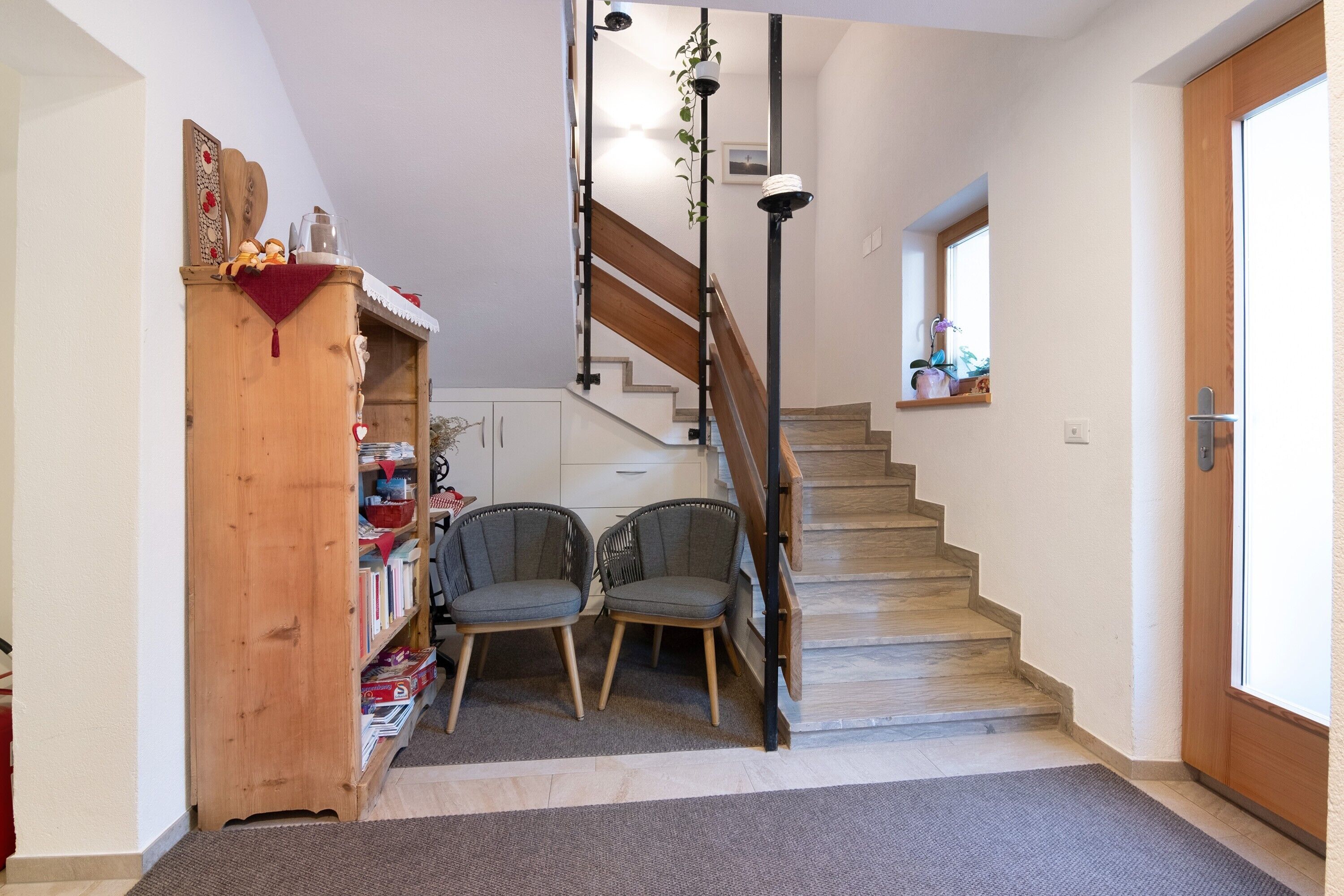A staircase leading to a second floor, a wooden cabinet with books, two chairs, and a small window with a view.