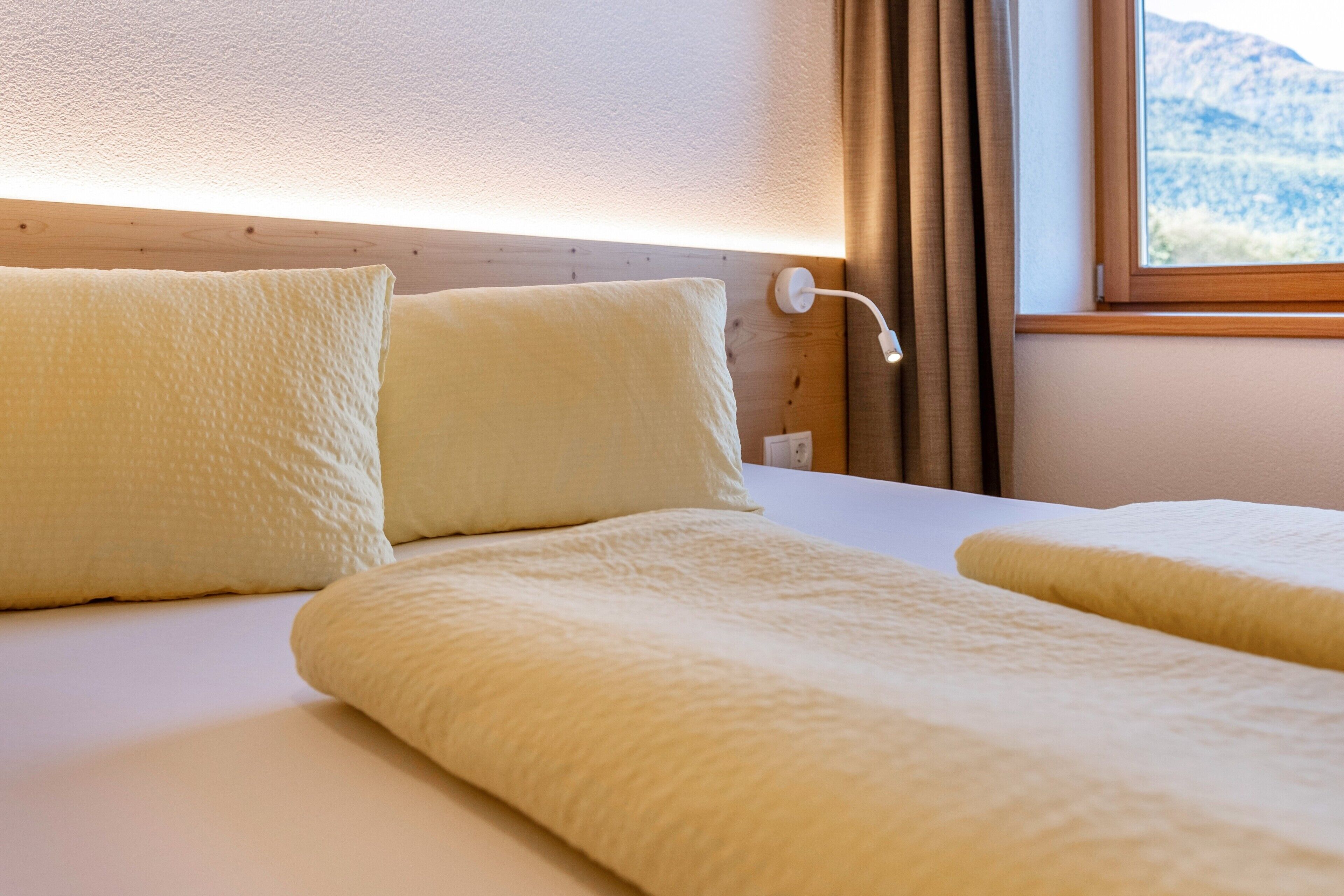 Habitación de hotel con cama, almohadas, una lámpara de noche y una ventana con vistas a las montañas.