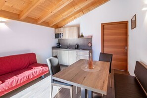 A compact kitchen with a microwave, coffee maker, and kettle, a dining table with chairs, and a red sofa.