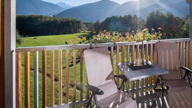 A balcony with a table and chairs, overlooking a green field and mountains.
