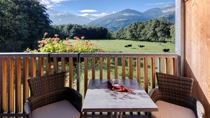 Ein Balkon mit Korbmöbeln, einem Tisch mit einem Glas und Blick auf ein grünes Feld mit Kühen und Bergen.