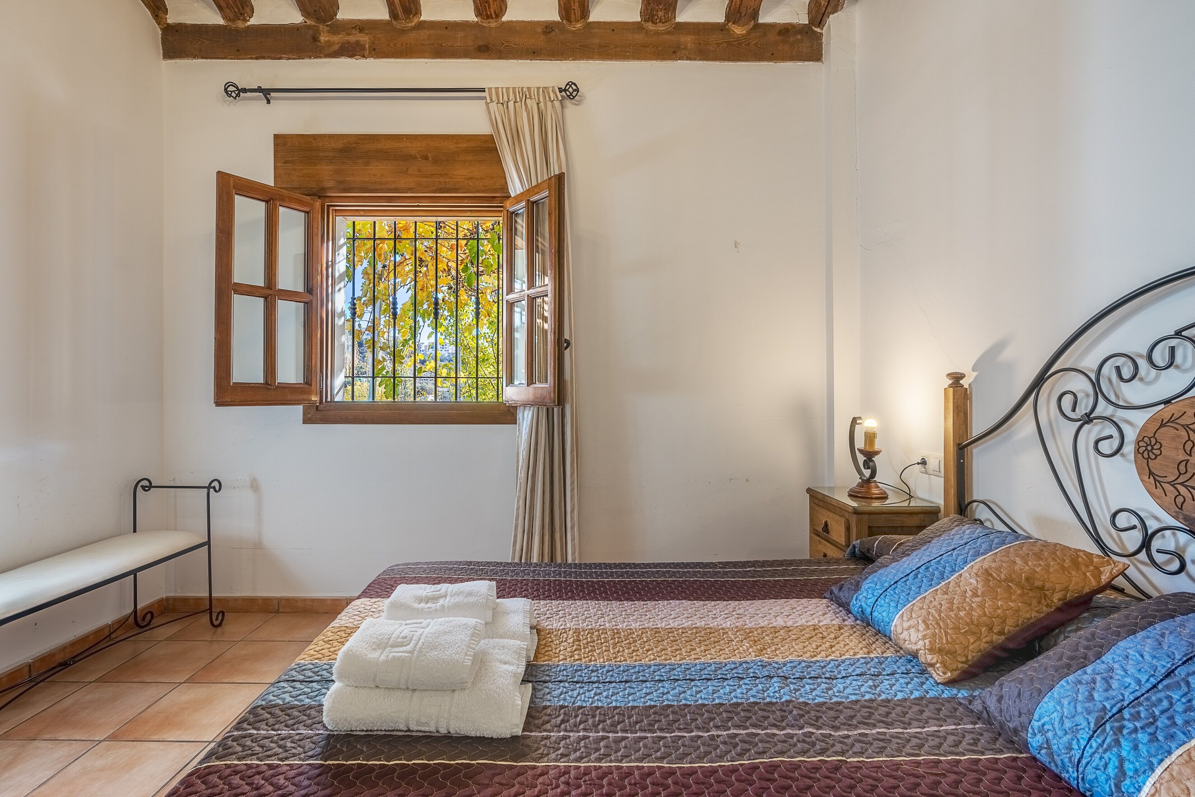 A bedroom with a bed, a bench, a window with curtains, and a nightstand with a lamp.