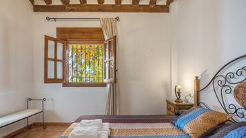 A bedroom with a bed, a bench, a window with curtains, and a nightstand with a lamp.
