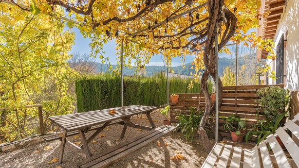 Een houten picknicktafel en een bank in een tuin met geel herfstblad en uitzicht op heuvels.