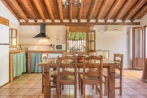 A rustic kitchen with wooden beams, a dining table with chairs, a window with curtains, and a microwave. - Apartment 'Casa Rural Cortijo El Plantio' with Mountain View, Wi-Fi and Air Conditioning (Granada)