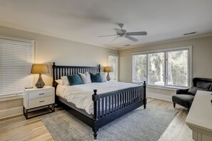 Une chambre à coucher comprenant un lit, deux tables de chevet, une commode, une chaise, un ventilateur de plafond et de grandes fenêtres.