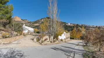 Un camí rural que condueix a un edifici blanc amb mur de pedra, envoltat d'arbres i un turó al fons.