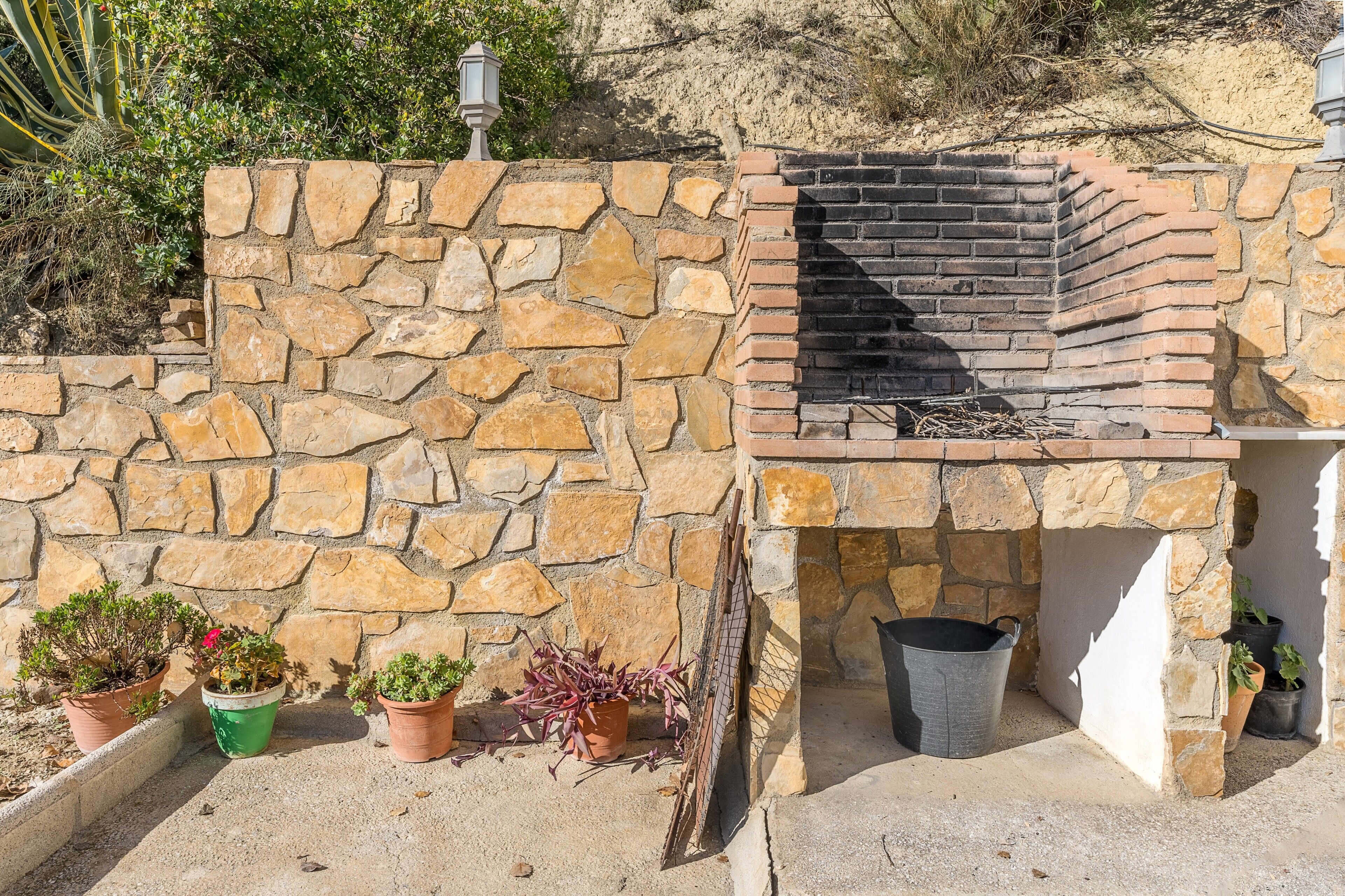 A stone wall with a brick oven, potted plants, and a metal grill.