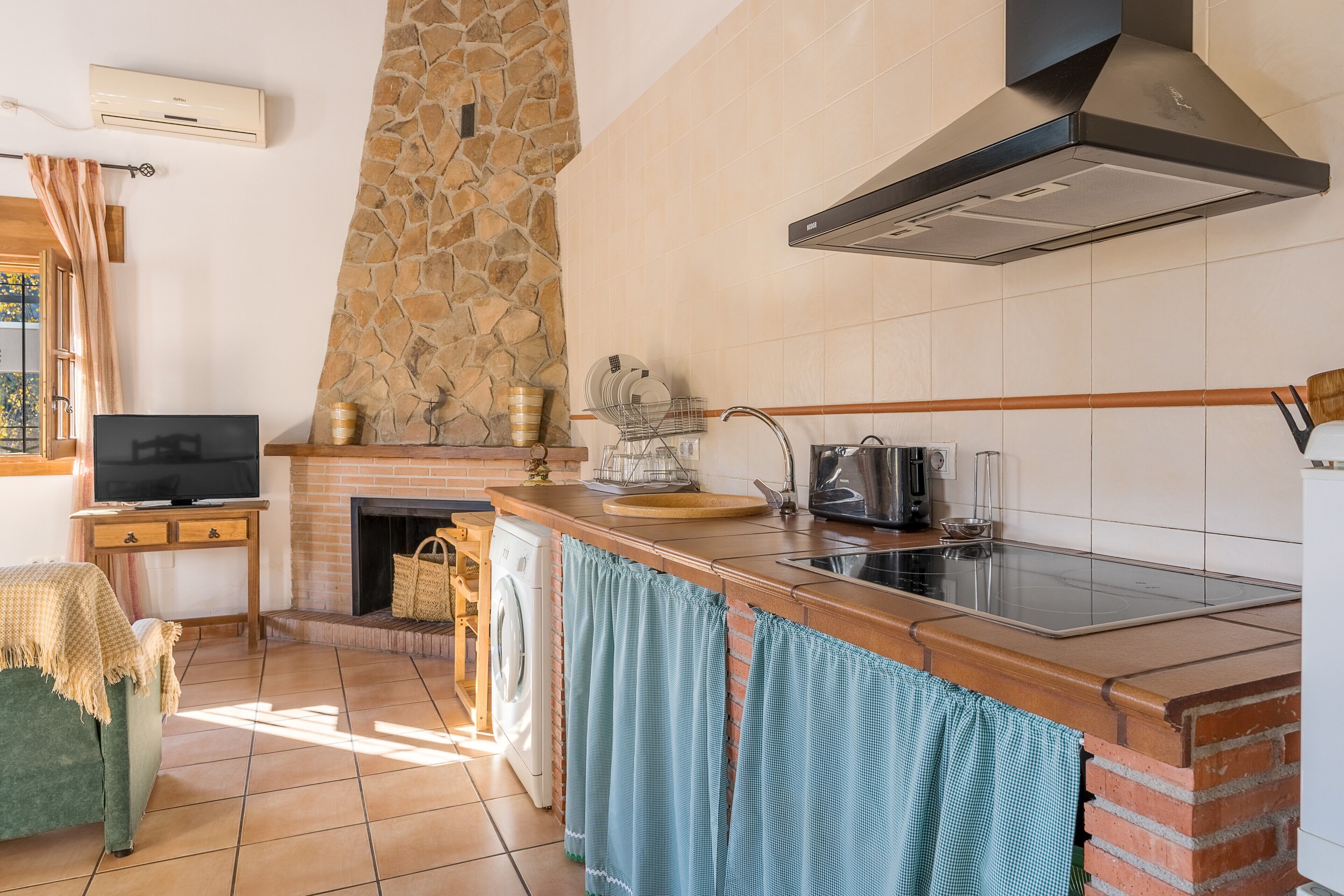 Una cocina con chimenea de piedra, televisor de pantalla plana, lavadora y un ventanal grande.