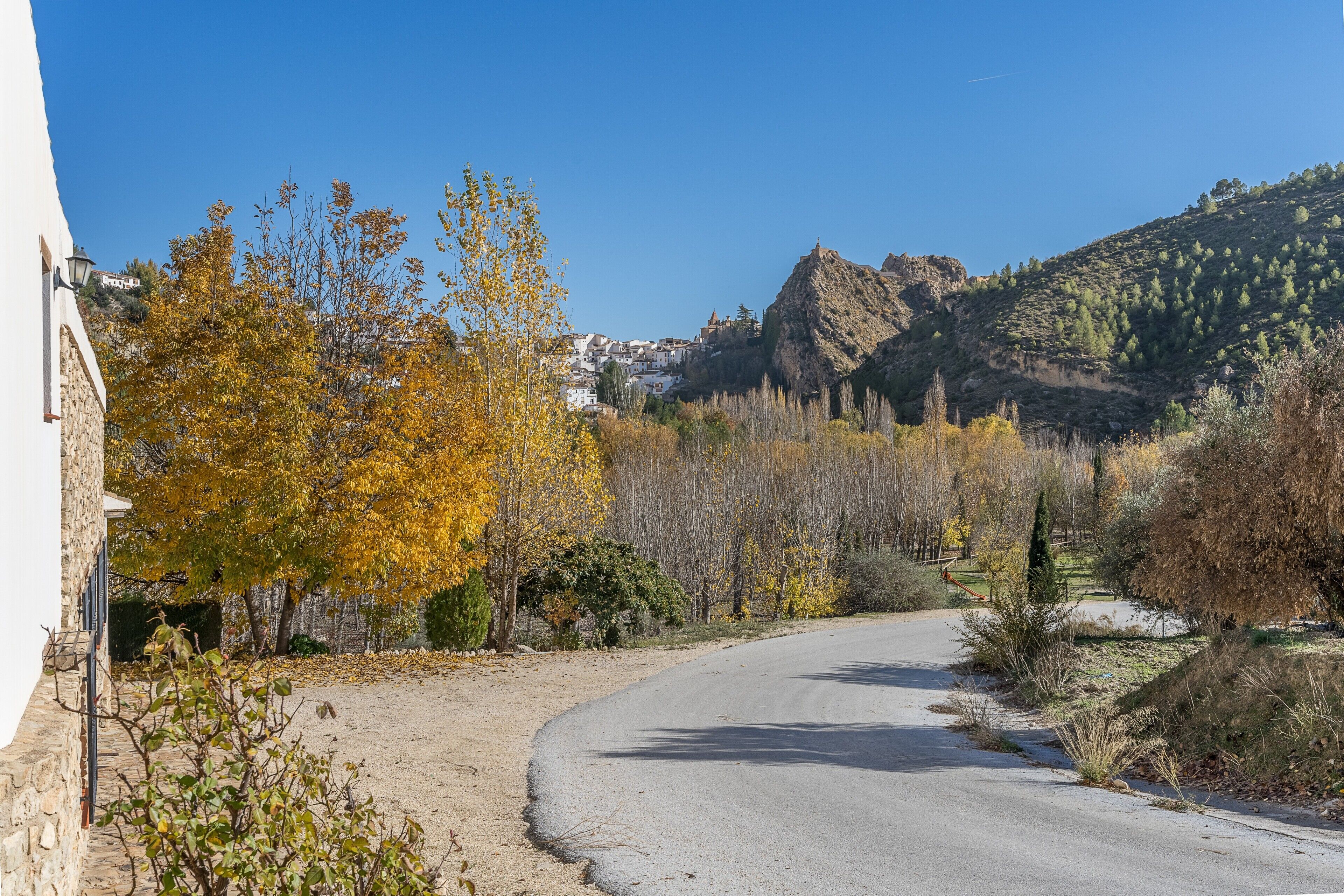 Una carretera serpenteante con un edificio de piedra a la izquierda, árboles con follaje otoñal y una colina con un afloramiento rocoso al fondo.