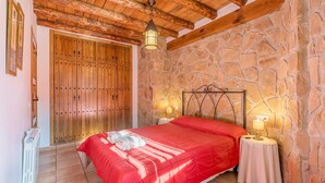 Un dormitorio con pared de piedra, puerta de madera, cama con colcha roja, dos lámparas de noche y un radiador.