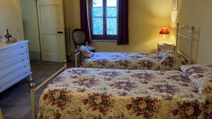 Ein Schlafzimmer mit zwei Betten, einem Schminktisch, einem Stuhl, einem Fenster mit Vorhängen, einer Lampe und einem gerahmten Bild an der Wand.