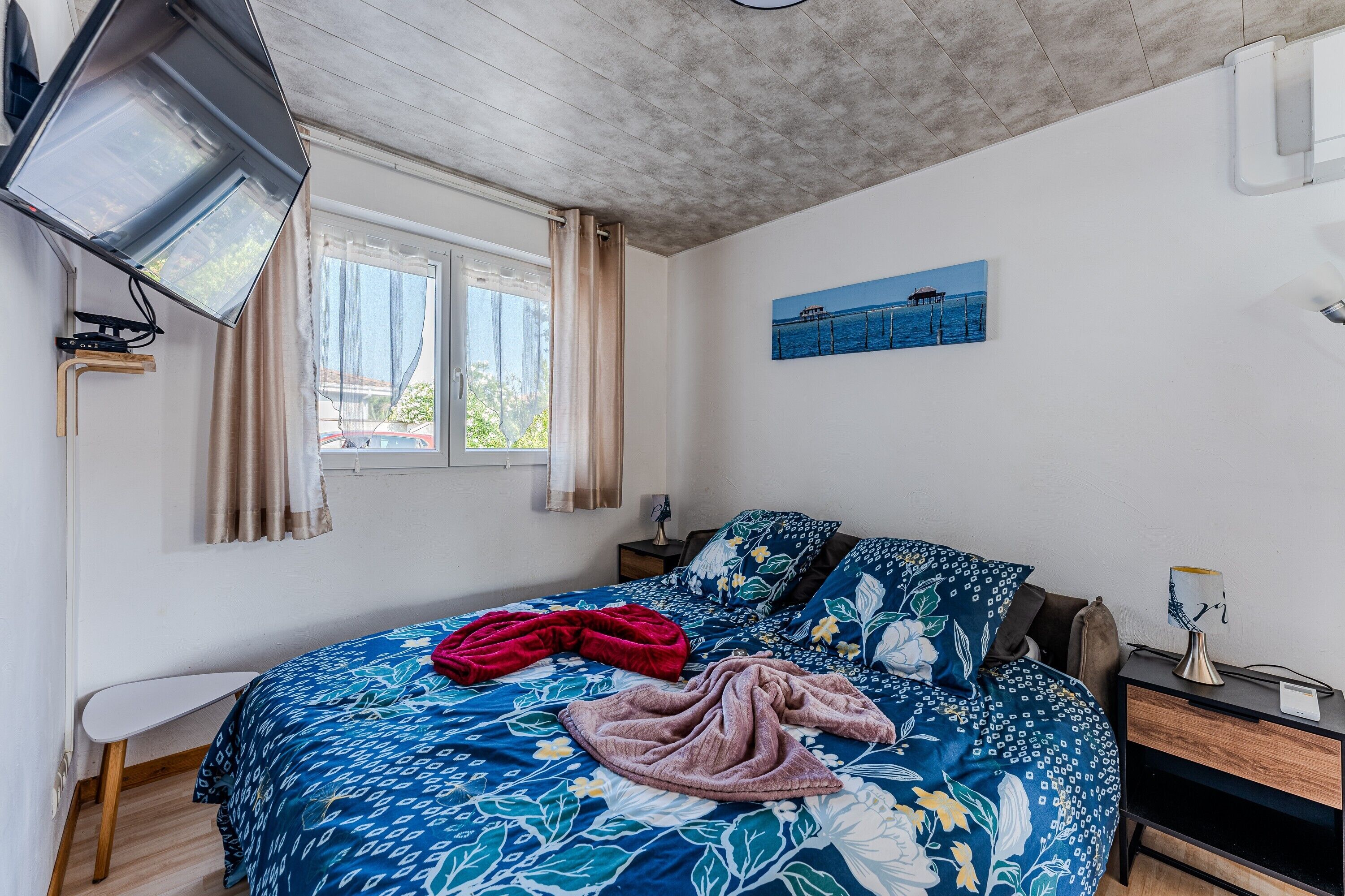 Een slaapkamer met een bed, een nachtkastje, een raam met gordijnen, een schilderij aan de muur en een plafondventilator.