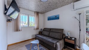 Une pièce comprenant un canapé, une petite table, un téléviseur fixé au mur, une fenêtre avec des rideaux et un tableau accroché au mur.