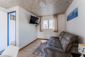 Ein Wohnzimmer mit einer Couch, einem an der Wand montierten Fernseher, einem kleinen Tisch und einem Fenster mit Vorhängen.