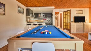 Una mesa de billar con bolas, un televisor y un techo de madera en una habitación con una cocina al fondo.