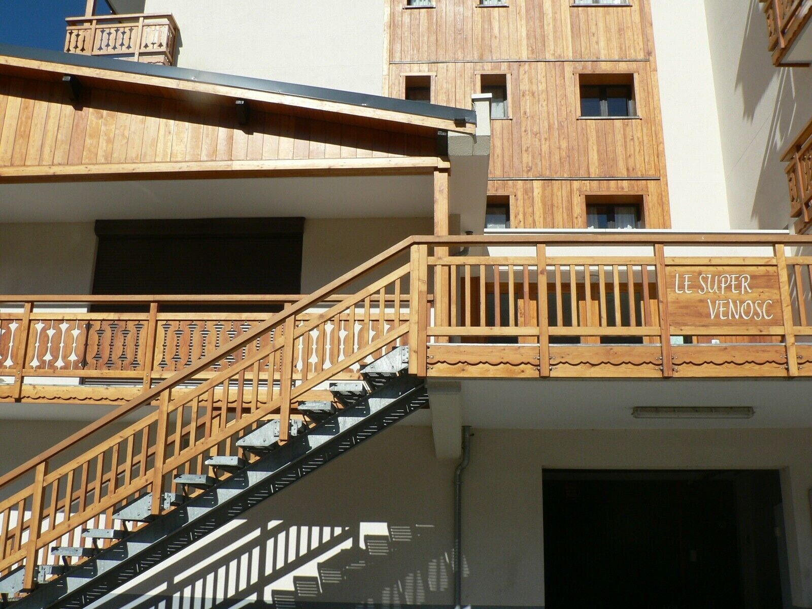 Un escalier en bois avec une rampe, menant à un bâtiment doté de nombreuses fenêtres et d’une enseigne sur laquelle on peut lire « LE SUPER VENOSC ».