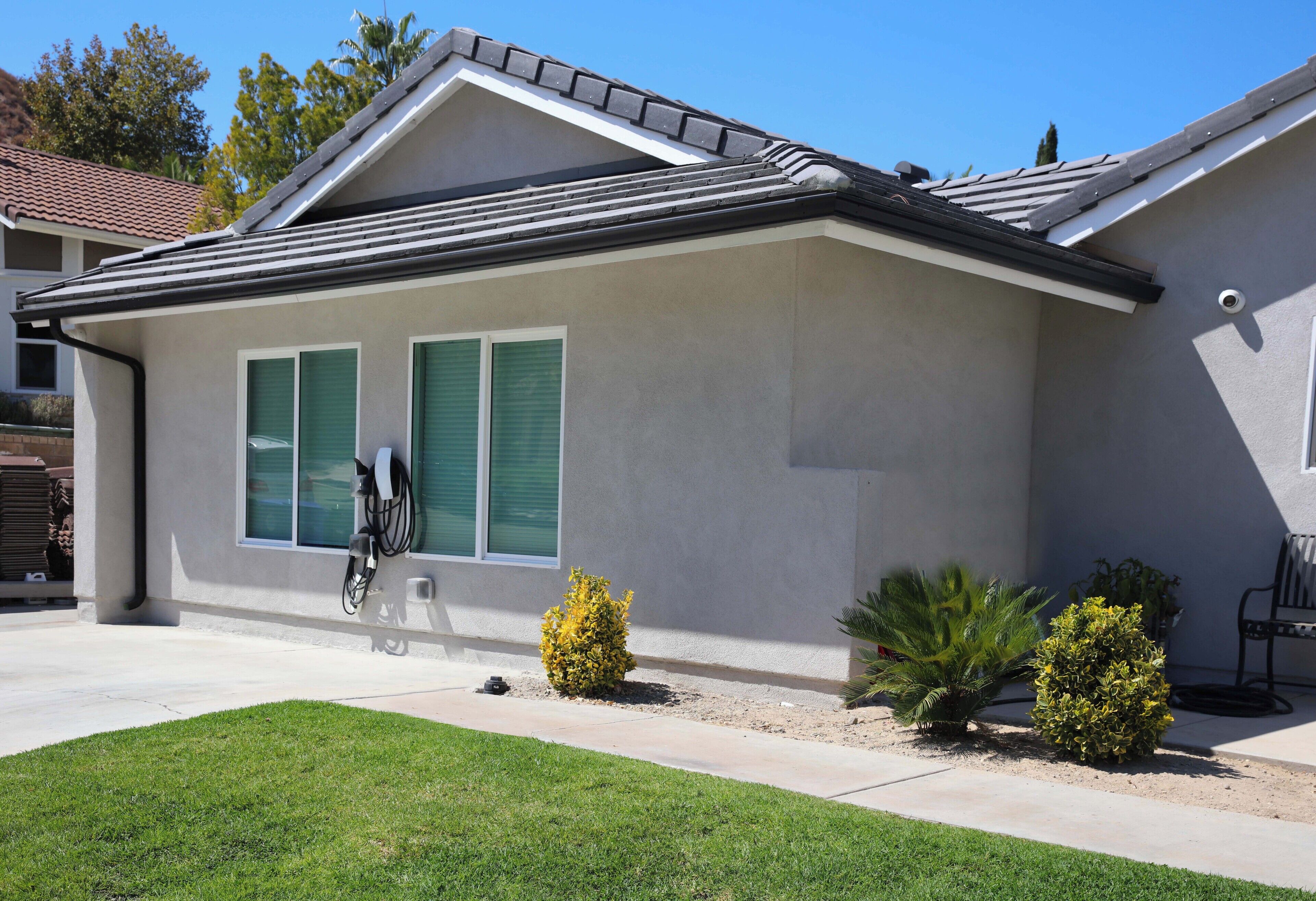 A single-story house with a grey roof, green shutters, and a small front yard with two bushes.