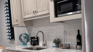 A modern kitchen with light wood cabinets, a stainless steel dishwasher, a microwave, and a wine rack with bottles.