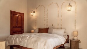 A bedroom with a wooden bed, a round nightstand, a wall-mounted lamp, and a door.