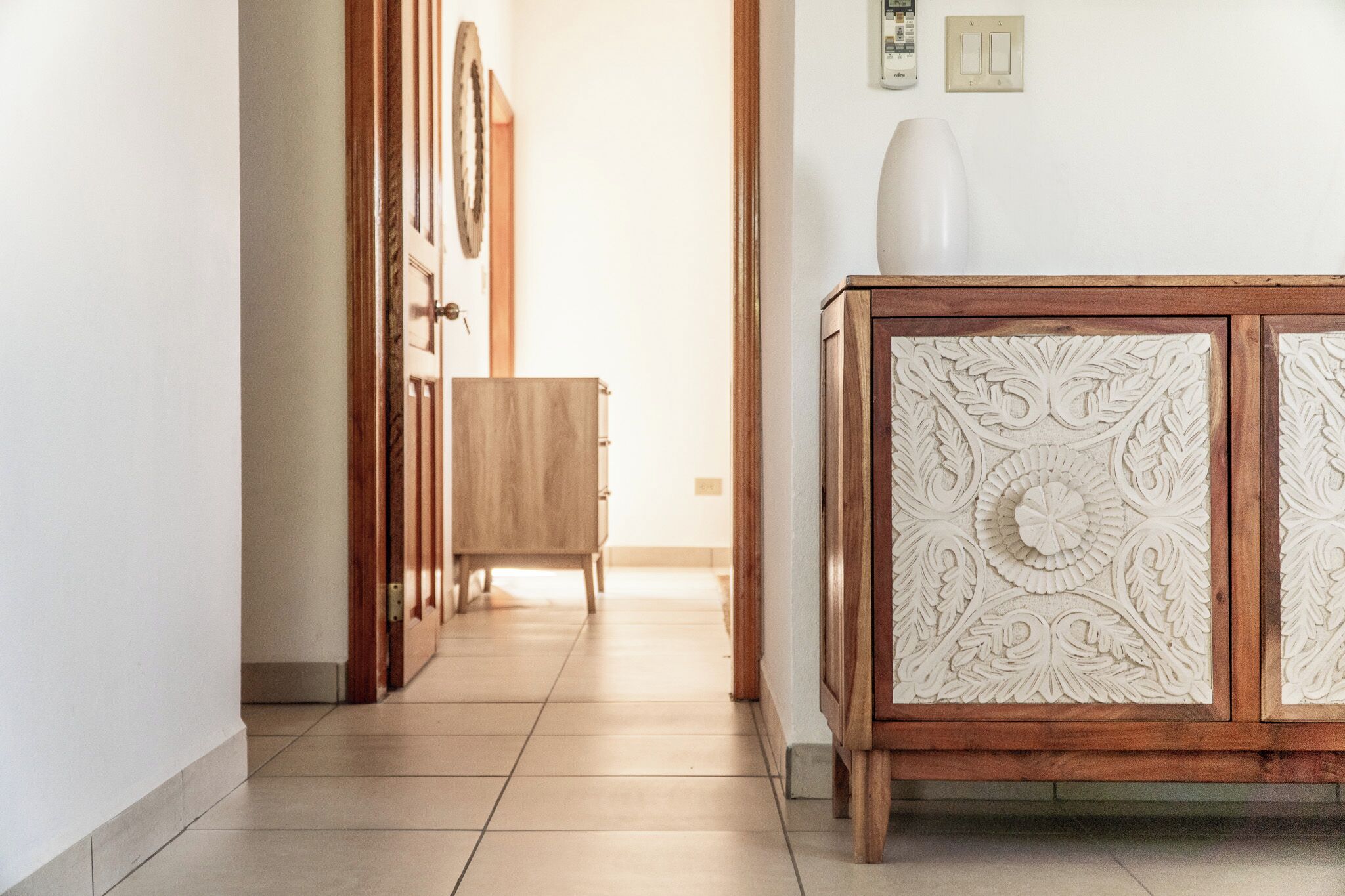 A hallway with a wooden cabinet featuring intricate carvings, a door with a decorative handle, and a light switch on the wall.