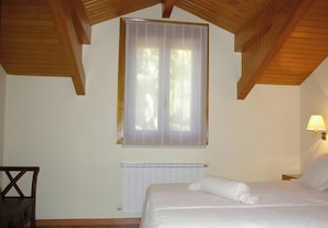 Une chambre avec un plafond en bois, une fenêtre avec des stores, un radiateur, un lit avec une literie blanche et une table de chevet avec une lampe.