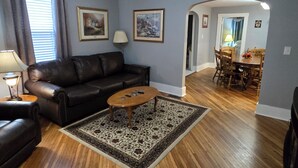 A living room with a black leather sofa, a wooden coffee table, and a patterned rug. There is a dining area with wooden chairs and a table in the background. - Charming 2-bedroom bungalow with WiFi and AC in serene Mount Carmel (Mount Carmel)