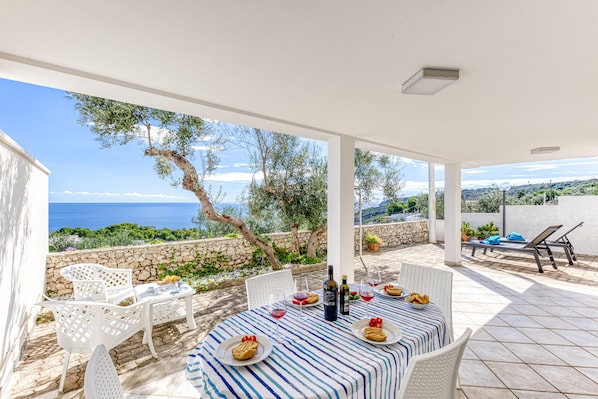 An outdoor patio with a table set for dining, chairs, and a view of the sea.