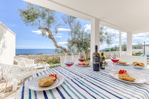 A table set with plates of food, glasses of red wine, and bottles of olive oil, overlooking a scenic coastal view.