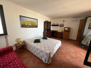 A bedroom with a bed, a nightstand, a lamp, a red sofa, a kitchen area with cabinets, a refrigerator, and a window.