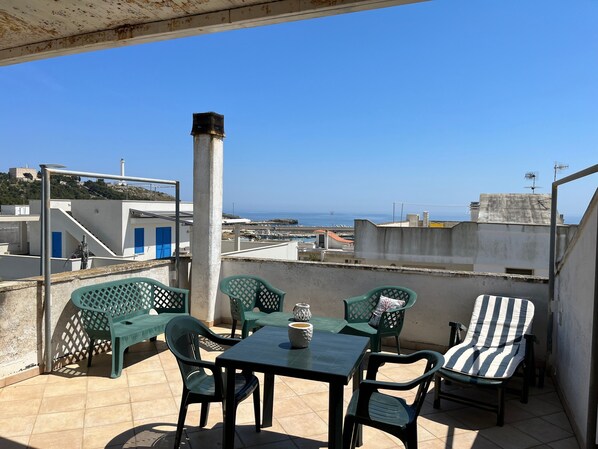 A rooftop terrace with a table and chairs, a view of the sea, and buildings in the background.