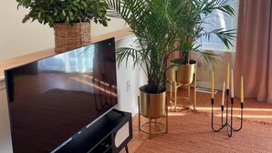 Een woonkamer met een flatscreen-televisie, een kamerplant, een vloerlamp en een raam met gordijnen.