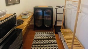 Een kleine kamer met een magnetron, een kast met glazen deuren, een houten plank, een kledingroede en een kamerkleed met dessin.