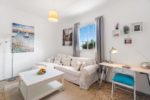 A white furnished room with a sofa, a desk, a chair, a lamp, a window with curtains, and wall art.