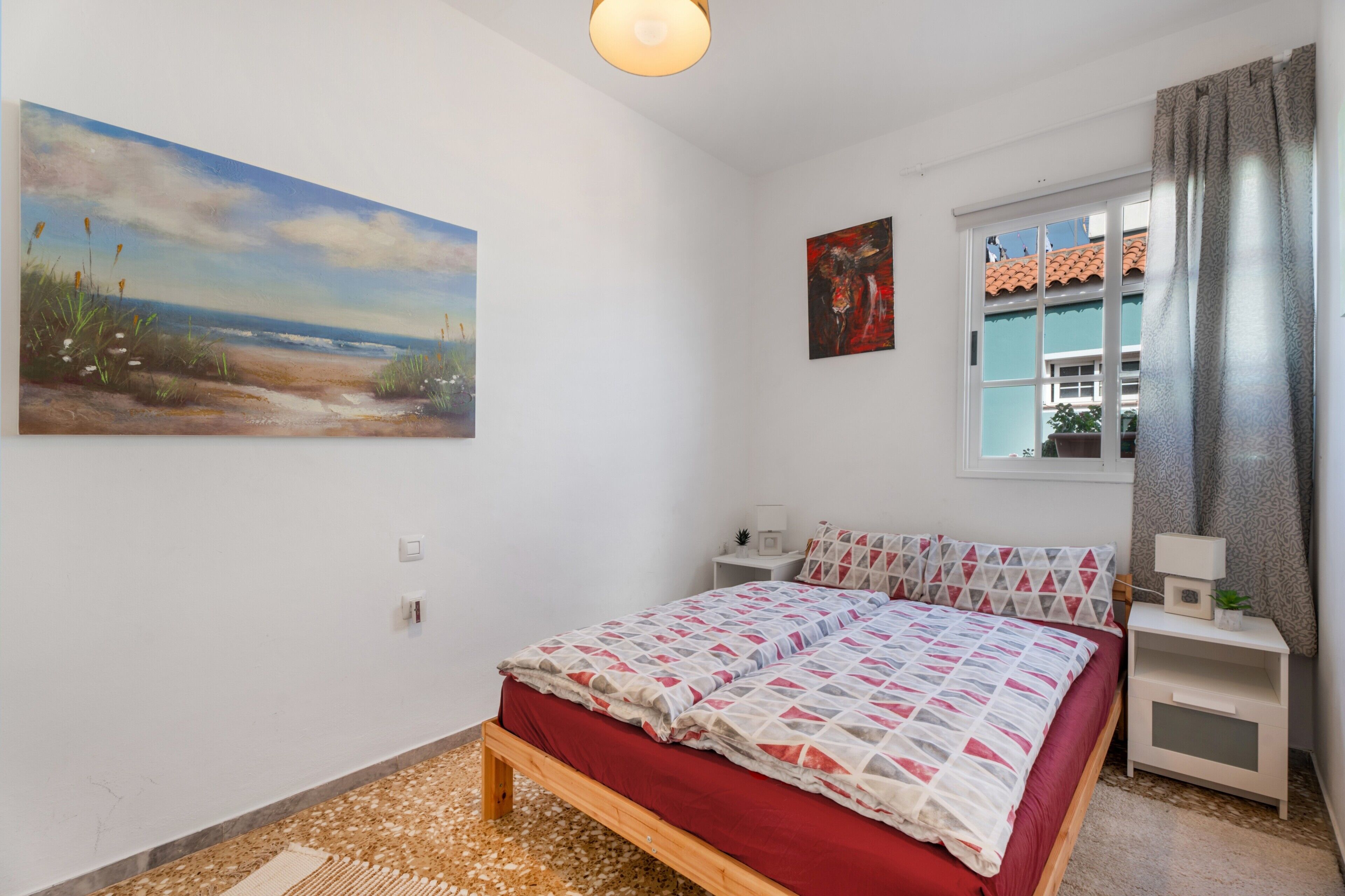 Een slaapkamer met een bed, nachtkastje, raam met gordijnen en een schilderij aan de muur.