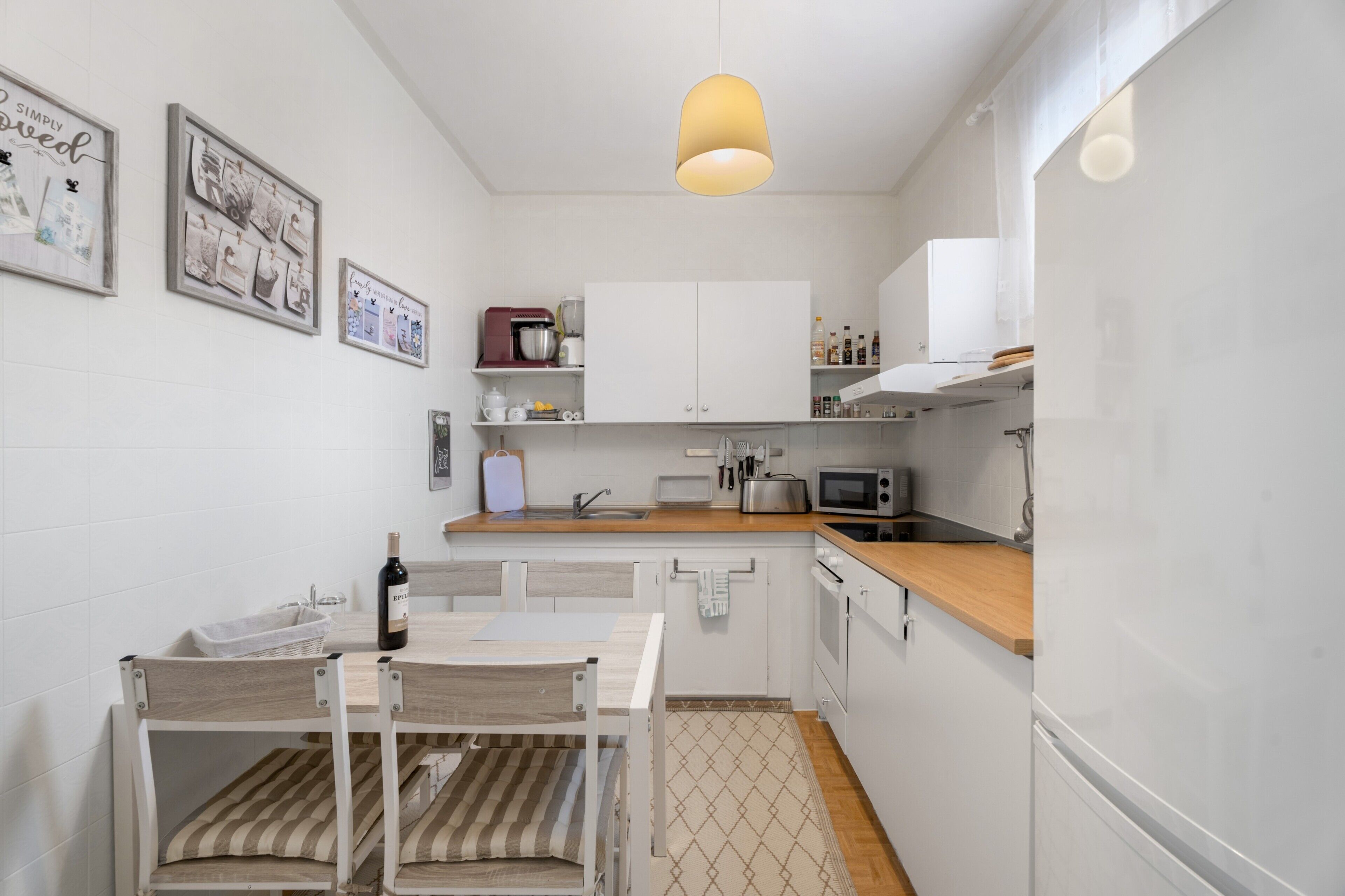 Una cocina compacta con armarios blancos, encimera de madera, lámpara colgante y comedor con mesa y sillas.