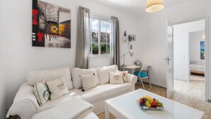 Een witte woonkamer met een bank, een bijzettafeltje met fruit, een bureau en een schilderij aan de muur.