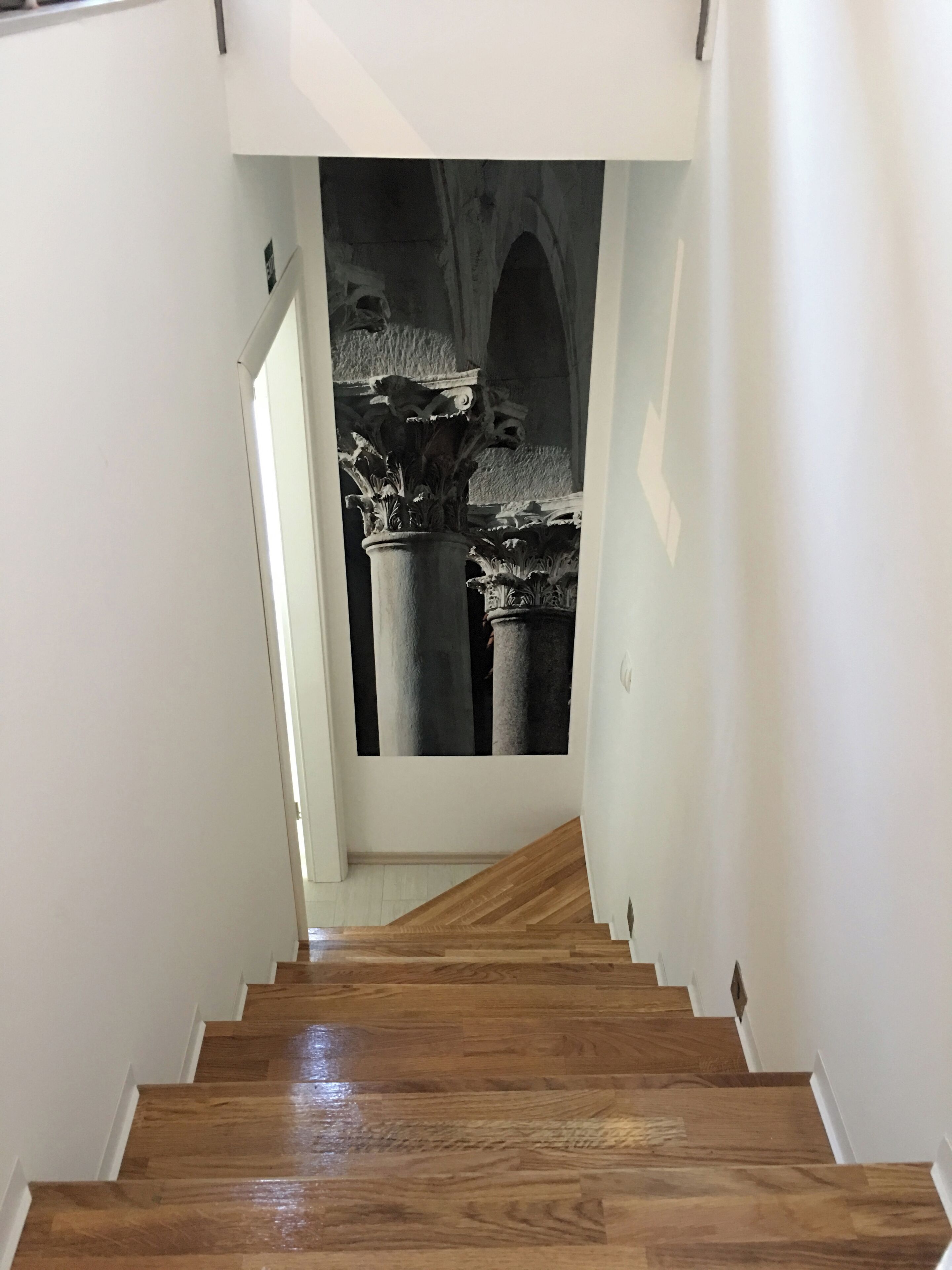 Uma escada de madeira leva a um quarto com uma fotografia grande em preto e branco de colunas e arcos antigos na parede.