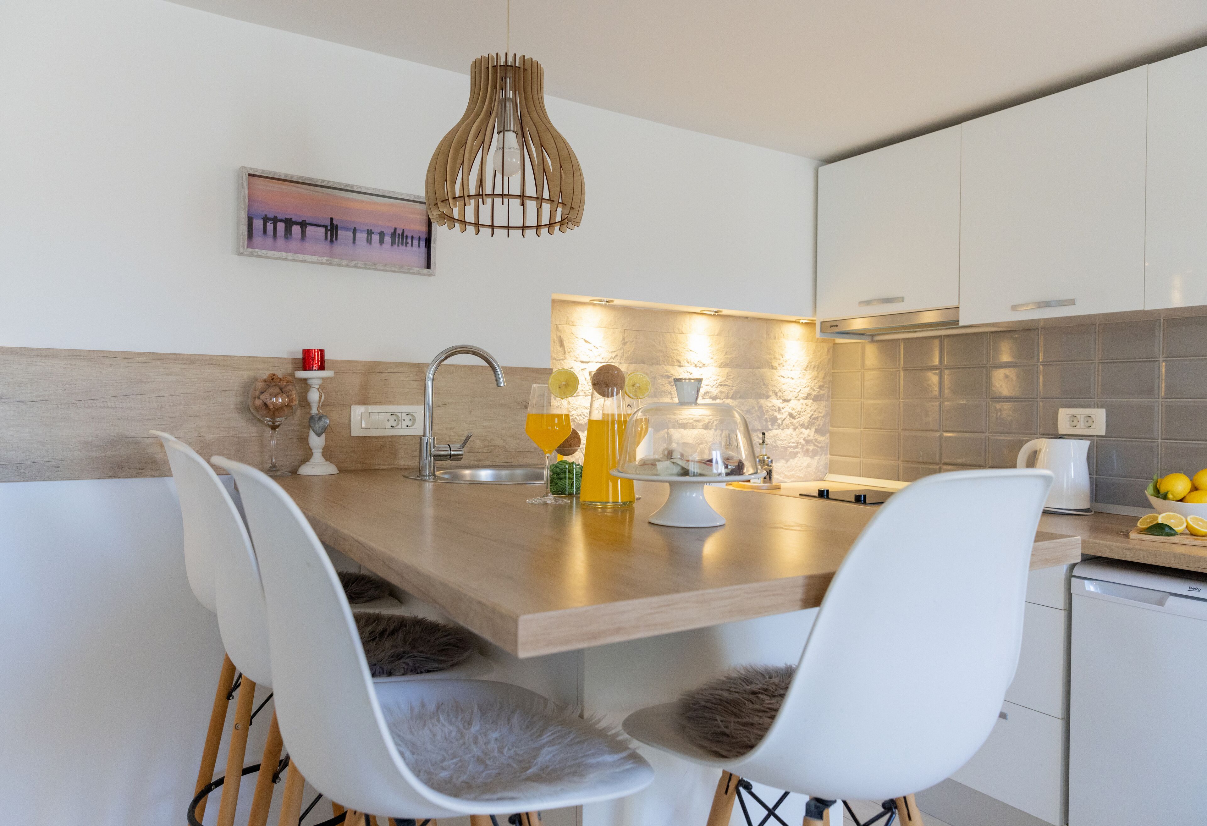 A modern kitchen with a wooden island, white chairs, a hanging light fixture, and a stone backsplash.
