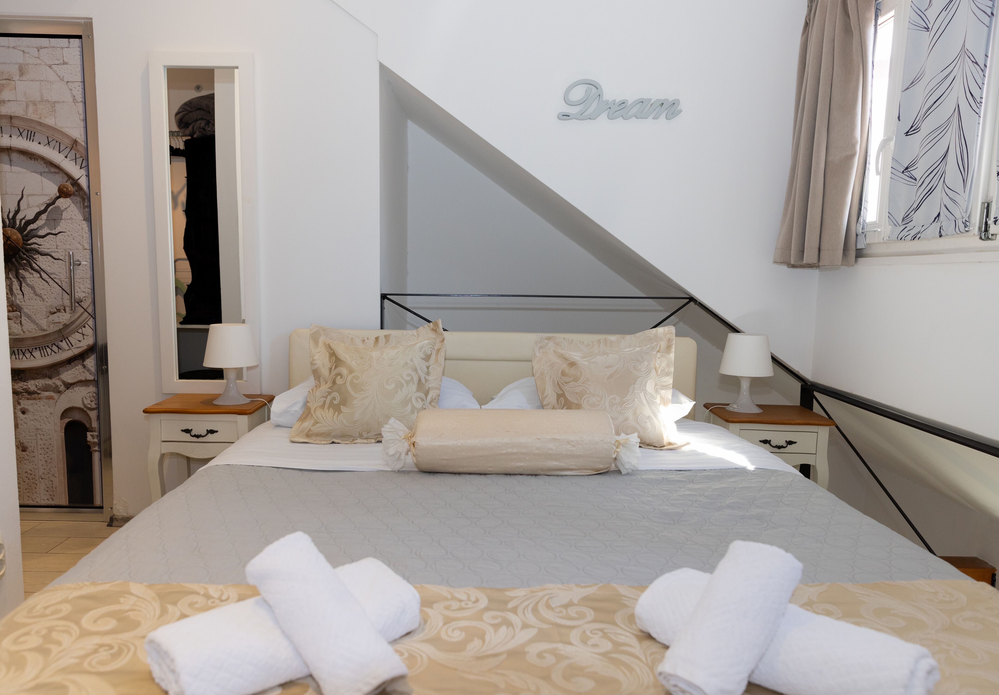 A neatly made bed with a headboard, two bedside tables with lamps, a large mirror, and a wall with the word "Dream" written on it.