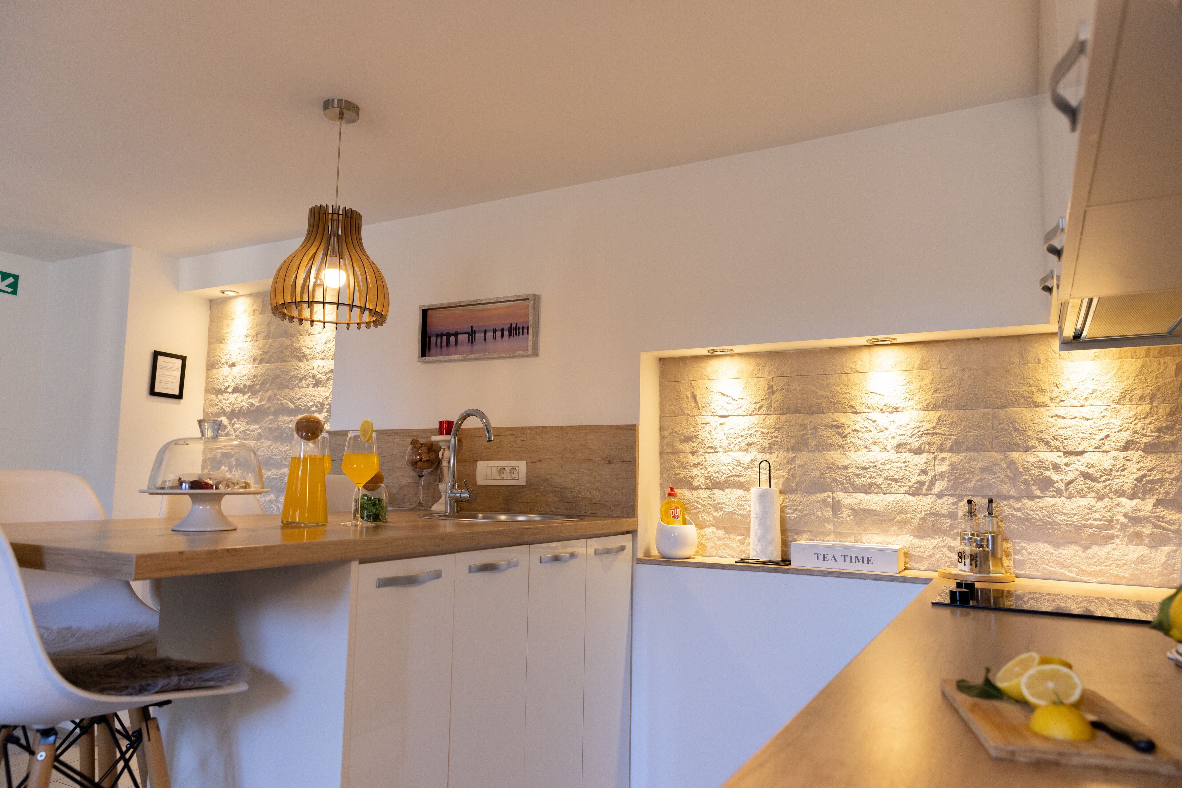 Une cuisine moderne avec un plan de travail en bois, un luminaire suspendu, un évier et une enseigne indiquant « HEURE DU THÉ ».
