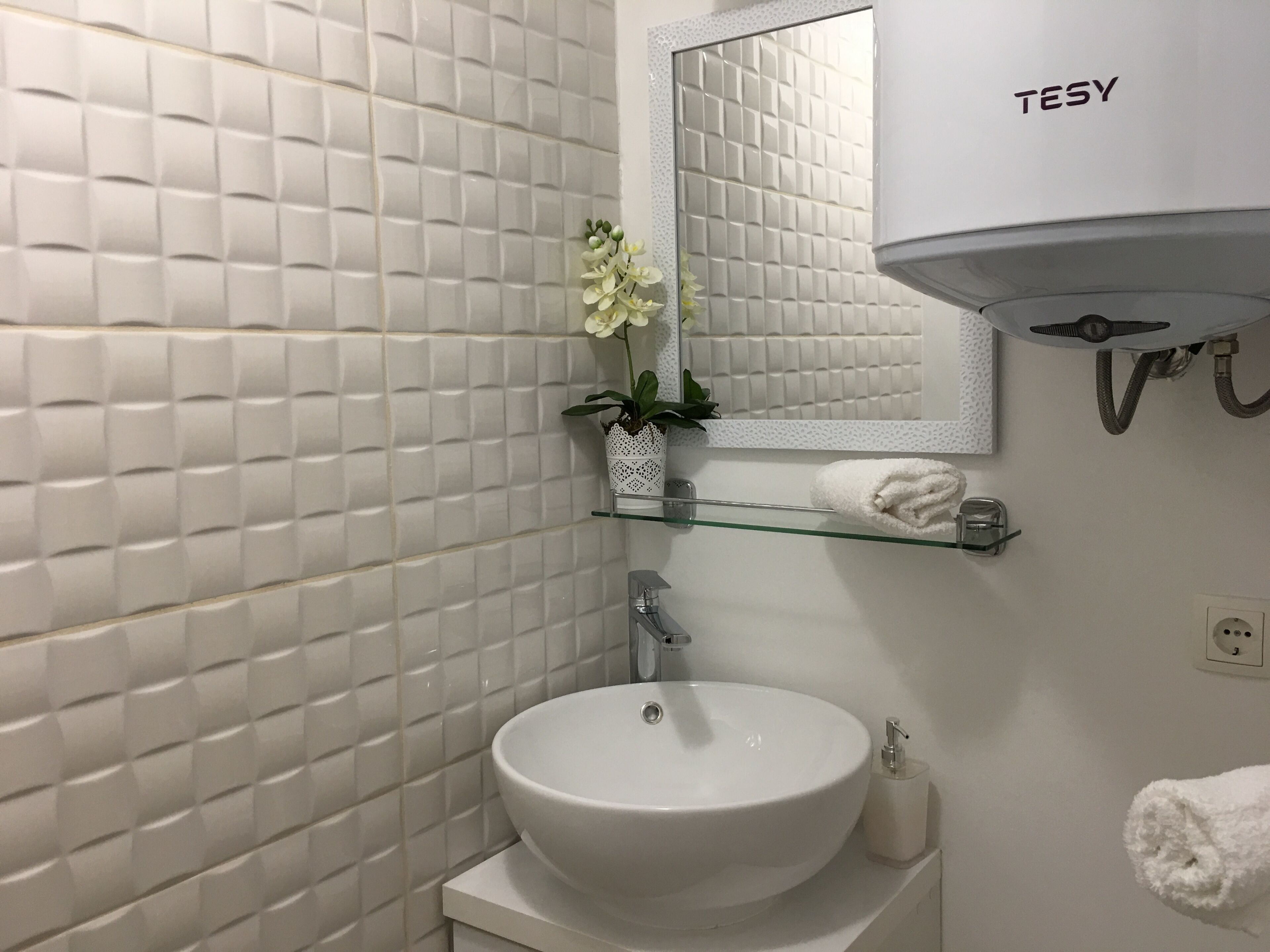 Een badkamer met een betegelde witte wand, een wit wastafelmeubel, een spiegel, een handdoekhouder en een plant.