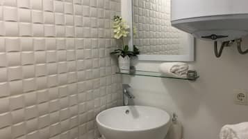 Een badkamer met een betegelde witte wand, een wit wastafelmeubel, een spiegel, een handdoekhouder en een plant.