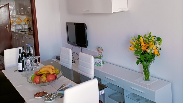 Un comedor moderno con una mesa blanca, sillas, una lámpara colgante rosa, un televisor de pantalla plana, un florero con flores y un ordenador portátil sobre la mesa.