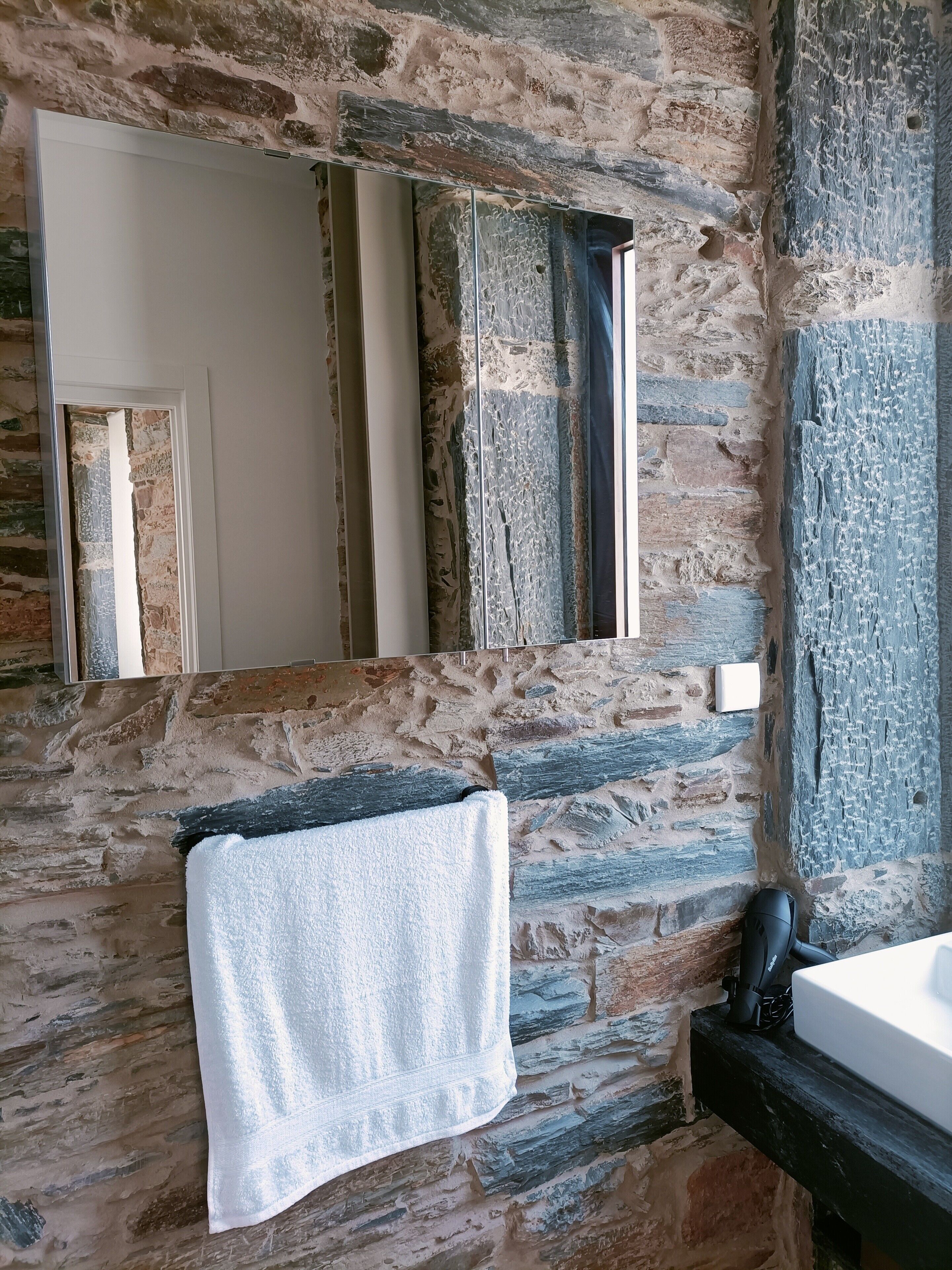 A bathroom with a stone wall, a mirror, a towel, and a black hairdryer.