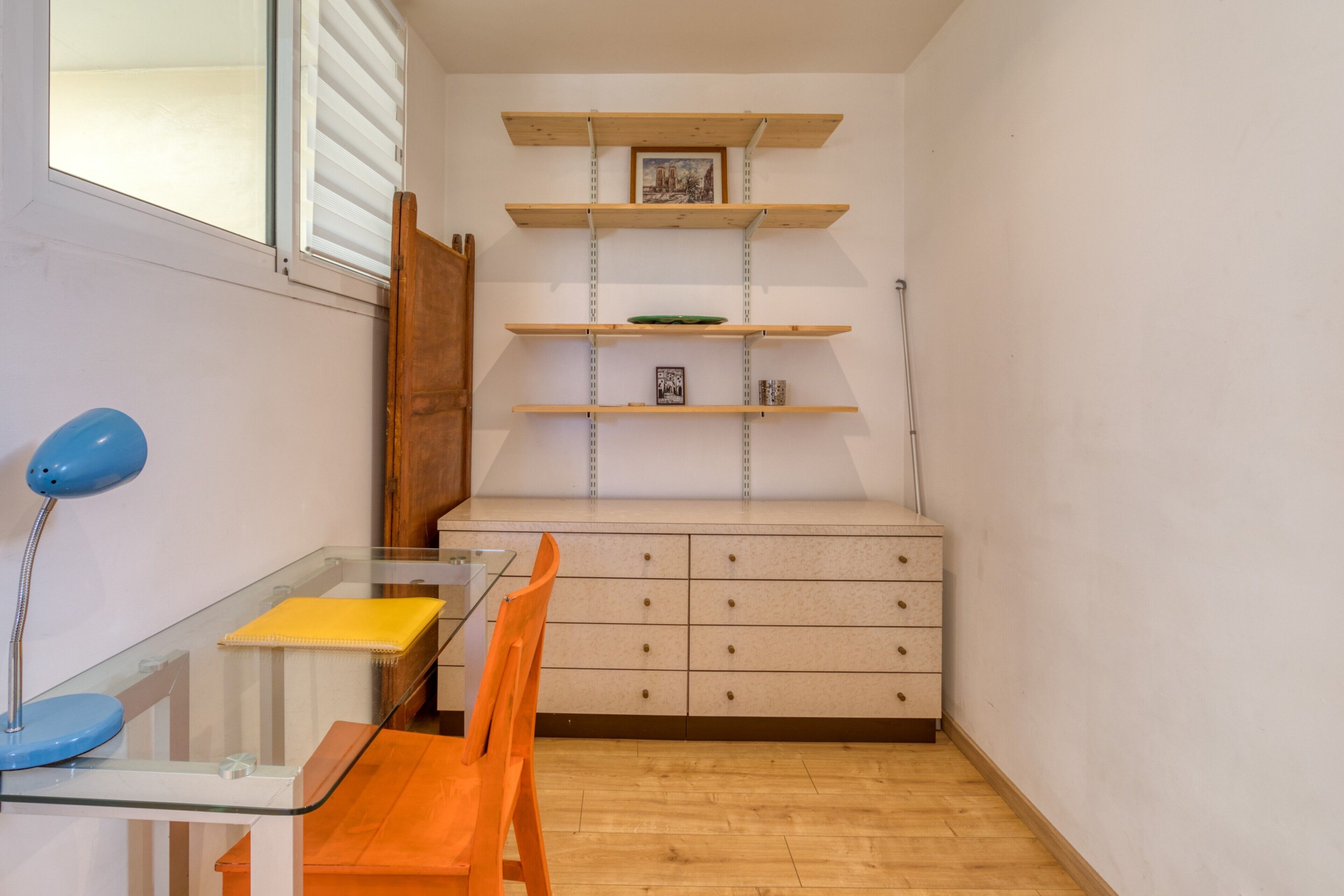 A room with a glass table, orange chair, wooden shelves, and a cabinet.