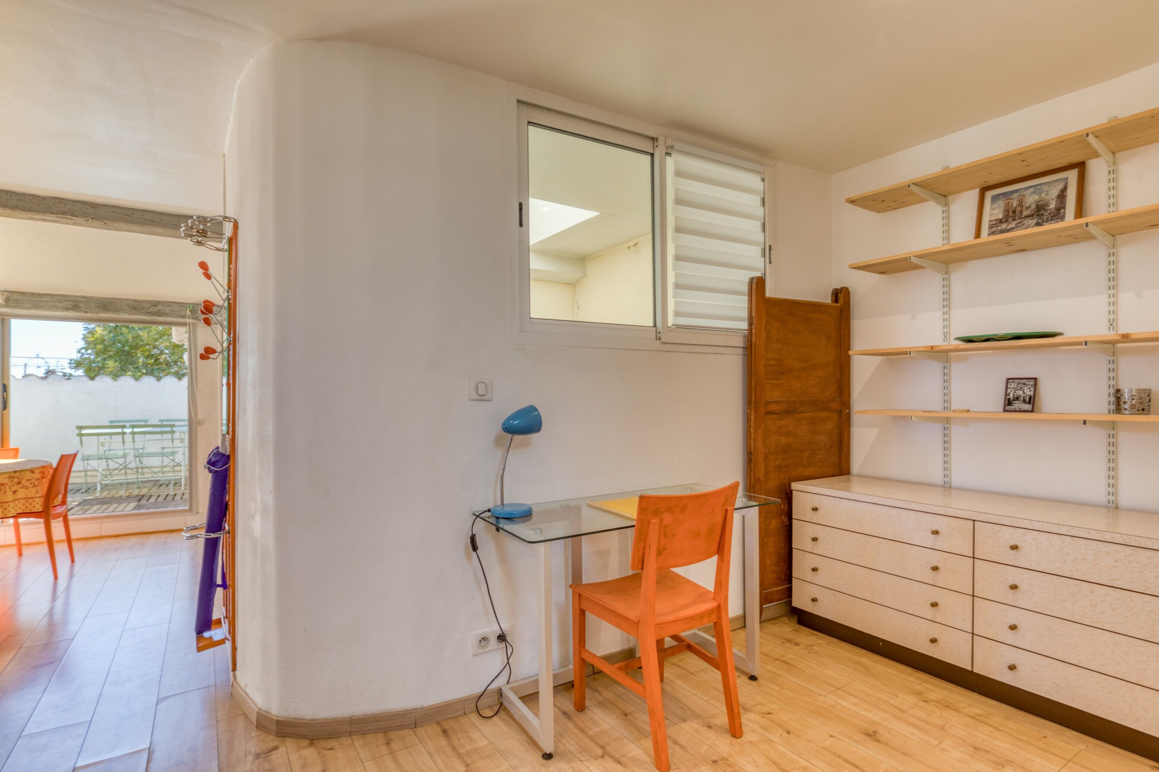 A room with a glass desk, an orange chair, a wooden cabinet, and a window with blinds.