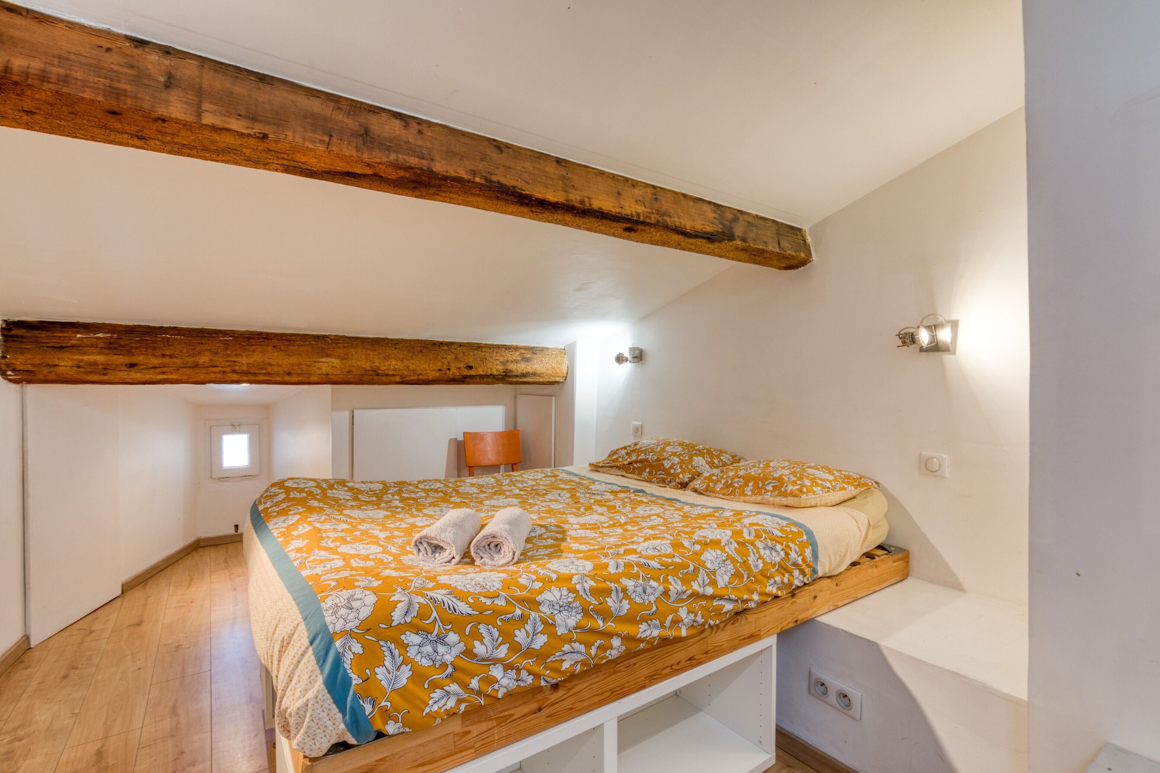 Een slaapkamer met een houten bedframe, een raam, een stoel en een wandlamp.