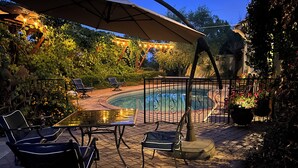 Un patio exterior con piscina, sillas de descanso y una pérgola con luces colgantes.